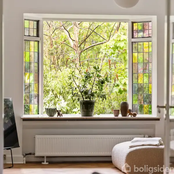 Stue med store vinduer med farvede glas, der viser udsigt til frodige træer udenfor. En plante står på vindueskarmen ved siden af dekorative genstande. En radiator er placeret under vinduet.