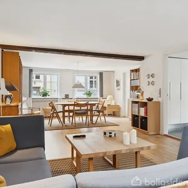 En moderne stue med en grå sofa og træbord, beliggende i et lyst rum. En spisestue med træstole ses i baggrunden ved siden af en stor vindue og reoler.
