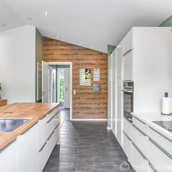 Køkken med moderne, hvide skabe og træbordplader, indrettet med vask og integrerede apparater. Rummet har flisegulv og trævæg med dør til have. Farverig kunst og grønne vægge.