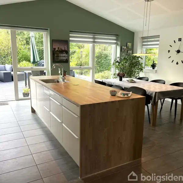 Køkkenø med vask står centralt i rummet, omgivet af klinkegulv og vinduer, der åbner ud til terrasse og grøn udsigt. Til højre ses et spisebord ved siden af en væg med et ur.