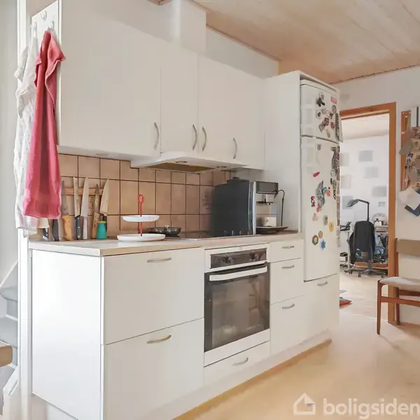 Et køkken med hvide skabe og en integreret ovn. En rød klud hænger ved siden af trapperne. I baggrunden ses et værelse med kontorstole. Køleskabet har magneter.