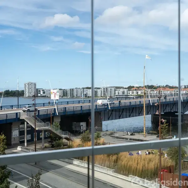 En hvid varevogn kører over en bro i vandkanten forbundet til en by med moderne boligblokke i baggrunden. Billede set gennem en glasvæg med refleksioner.