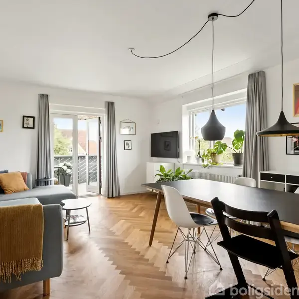 En stue med en grå sofa står ved siden af et spisebord og stole. Et stort vindue og en altandør giver lys, mens planter og kunst dekorerer væggene.