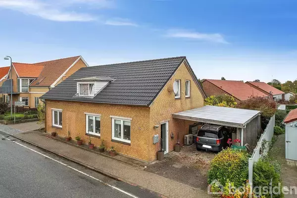 Et gult murstenshus med sort tag har en bil parkeret under en carport. Huset står i en villaområde med lignende huse langs en vej.