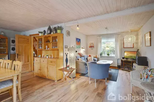 Stue med trægulv indeholder spisebord, stole, reol, og sofa med stole omkring sofabord. Et vindue fylder rummet med lys. Dekorationer pynter rundt omkring.