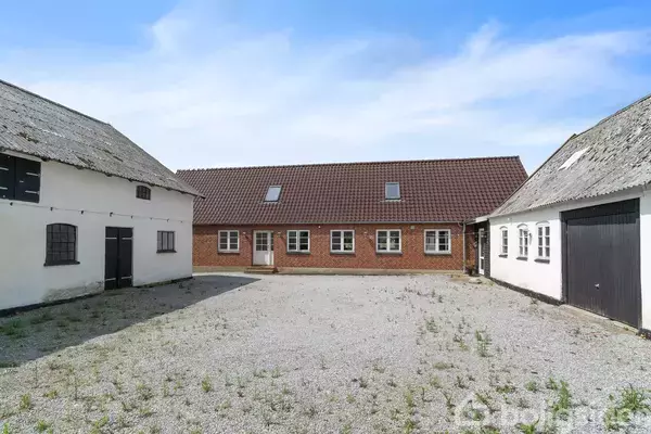 Et gårdanlæg med en central grusplads omgivet af tre bygninger; en rød murstensbygning med tagevinduer i midten og to hvidmalede længehuse på hver side.