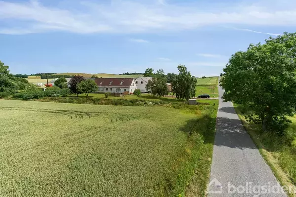 Et landsted med rødt tag ligger fredeligt omgivet af grønne marker og træer. En asfalteret vej fører op til ejendommen, der er placeret i et landligt område.