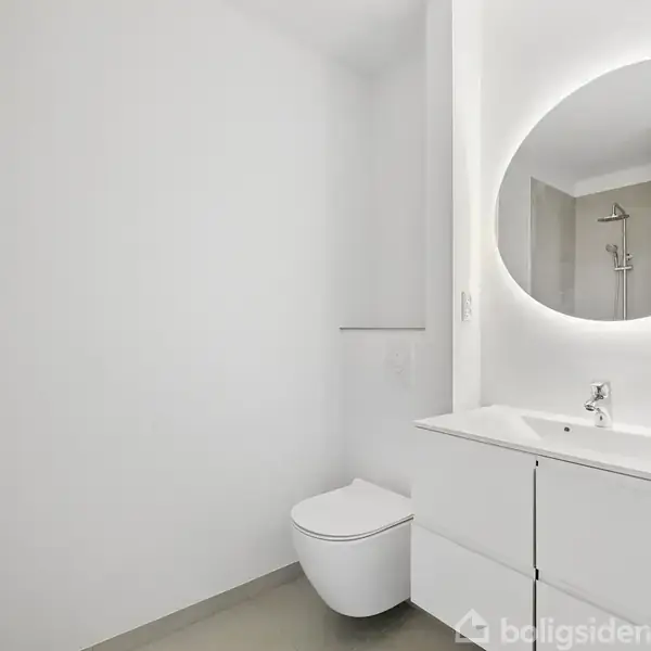 Toilet ved siden af en moderne vask med underskab, under et rundt spejl med baggrundsbelysning. Rummet er hvidt og minimalistisk med en bruser i baggrunden.
