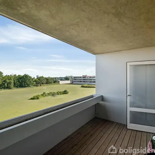 Balkon med trægulv, hvor døren står halvt åben. Den fører til en bolig, og udsigten viser en grøn park med træer og en bygning i baggrunden.