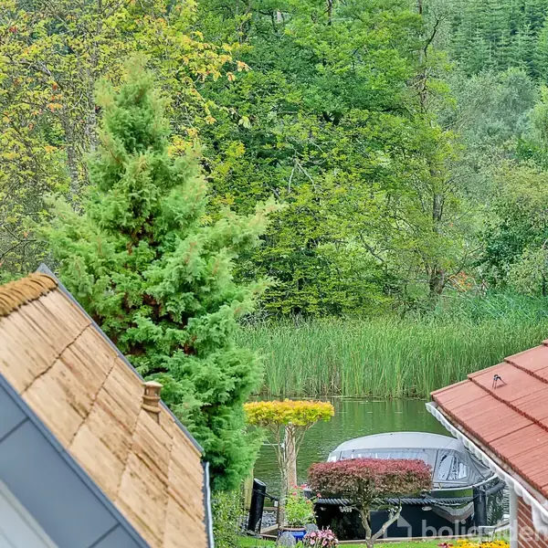 Tagbeklædte huse står stille blandt frodige træer og buske, med en sø i baggrunden omgivet af tæt siv. En båd ligger fortøjet nær vandkanten.