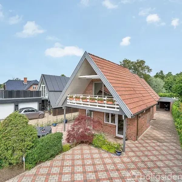 Et rødt murstenshus med et spidstag og balkon står ved siden af en brostensbelagt indkørsel. Omgivet af grønne buske og træer, er det placeret i et forstadsagtigt kvarter.
