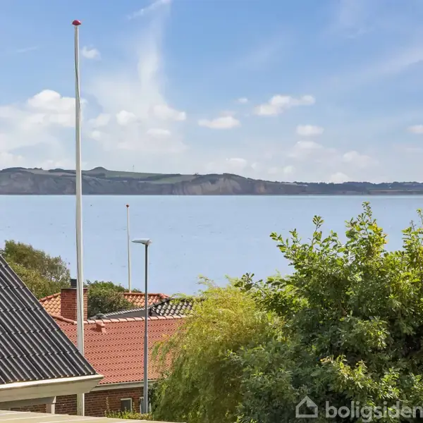 Skorsten står urørt på et hustag foran en rolig sø. I baggrunden ses en bakket skovhorisont, mens grønne træer omgiver bygningen og flagstangen.