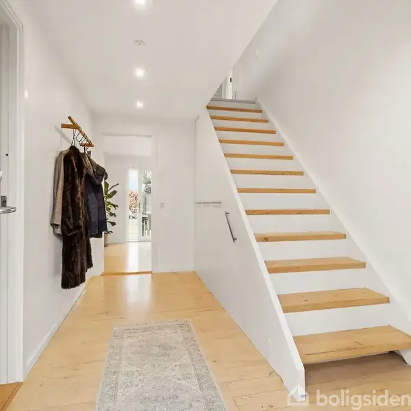 Trappe op ad en hvidmalet væg, flankeret af en række kroge med frakker, står i en lys gang med trægulv og tæppe, der fører ind mod et rum med planter.