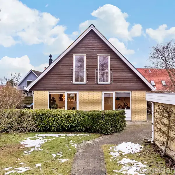 Et hus står i en have med en flisebelagt sti. Huset har en trekantet gavel og store vinduer. Der er snepletter på græsplænen og få buske omkring.