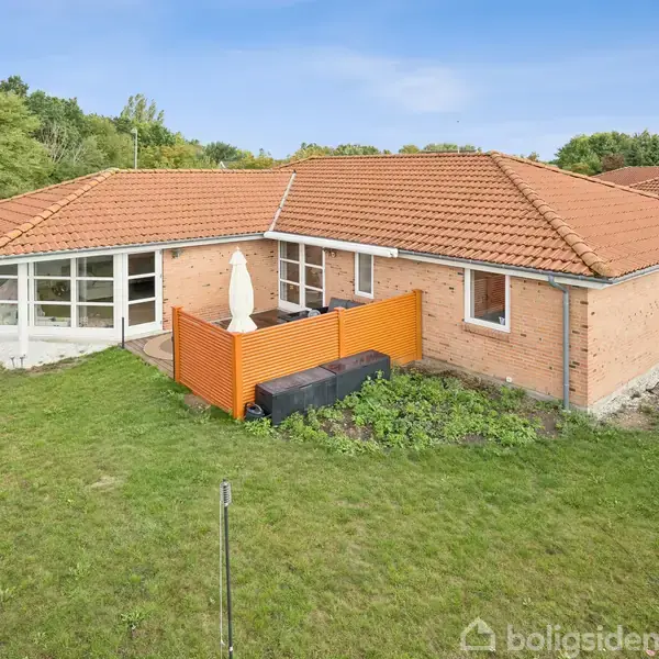 En enetages murstensvilla med rødt tegltag har en terrasse med en parasol og orange hegn omkring. Villaen er omgivet af en grøn have med træer i baggrunden.