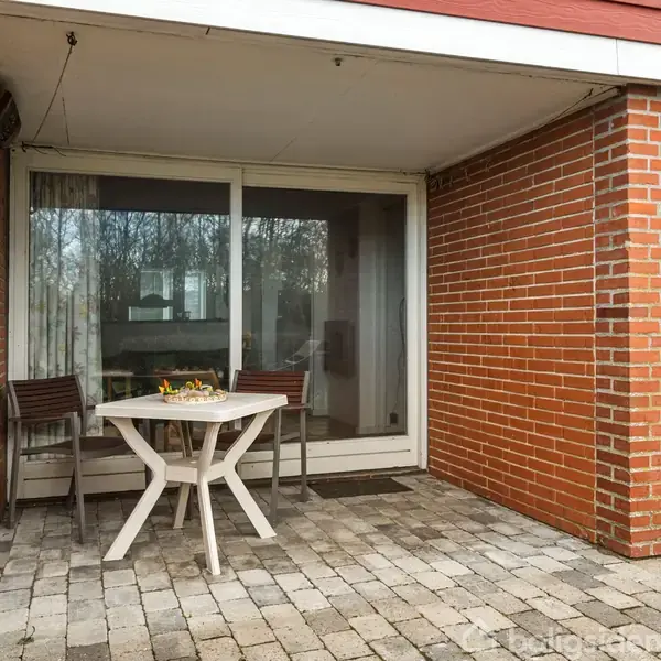 Et rundt bord med to stole står på en flisebelagt terrasse foran en murstensbygget altan med store glasskydedøre i et boligområde.