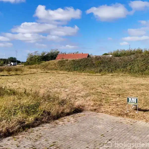 En græsmark ligger stille ved siden af en flisebelagt sti og et "Til salg" skilt. I baggrunden ses buske og et rødt tag under en blå himmel med skyer.