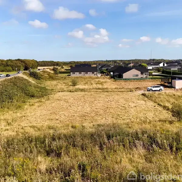 En åben græsmark med en bil parkeret ved en lille bygning på højre side. Omkringliggende huse og en vej til venstre under en klar himmel med få skyer.