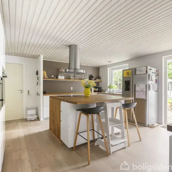 Et moderne køkken med en ø i midten, dekoreret med en gul vase med blomster. Køkkenet har hvide skabe, en stor køleskab med billeder, lyse vægge, og et vindue med udsigt til haven.