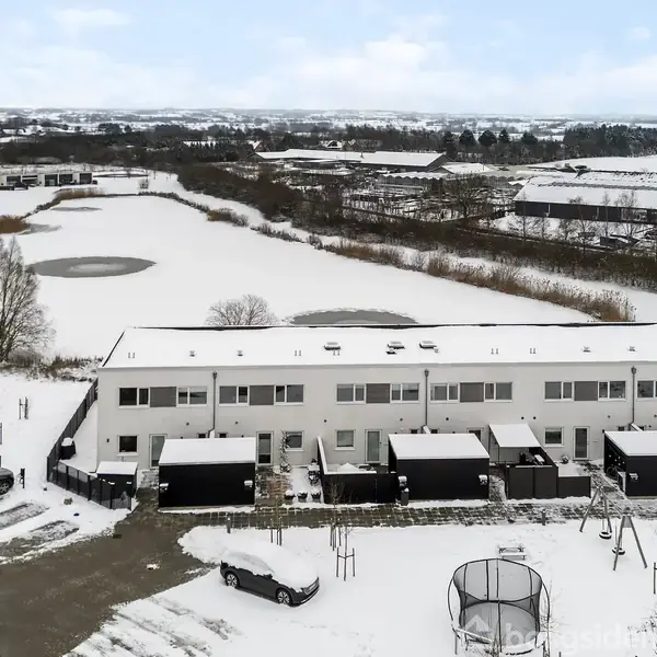 En række af to-etagers huse står stille i et snedækket landskab. Foran husene er flere parkerede biler. Omkring dem ses et åbent område med marker og en fjern bysilhuet.