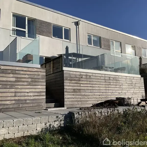 Lang rækkehus med store vinduer og glasbalkon. Træterrasser og niveauer skaber højdevariationer. Foran ses en mur af mursten og beplantning i sollys.