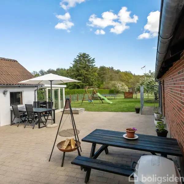 Terrassemøbler står på en flisebelagt gårdhave med et bord, stole og en parasol. I baggrunden ses en grøn græsplæne med en gyngestativ og omgivelser af træer.