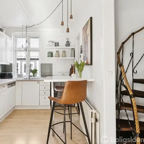 Spiraltrappe i mørkt metal ved siden af et moderne køkken med hvide skabe. Højbord med brun læderstol foran, og hængende lamper fra loftet.