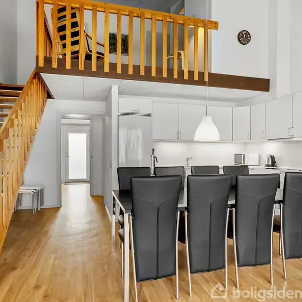 En spisestue med et sort spisebord og seks stole står i midten. Ovenpå er en trætrappe, der fører til en hems. Køkkenet til højre har hvide skabe og en moderne lampe. En ur hænger på væggen.