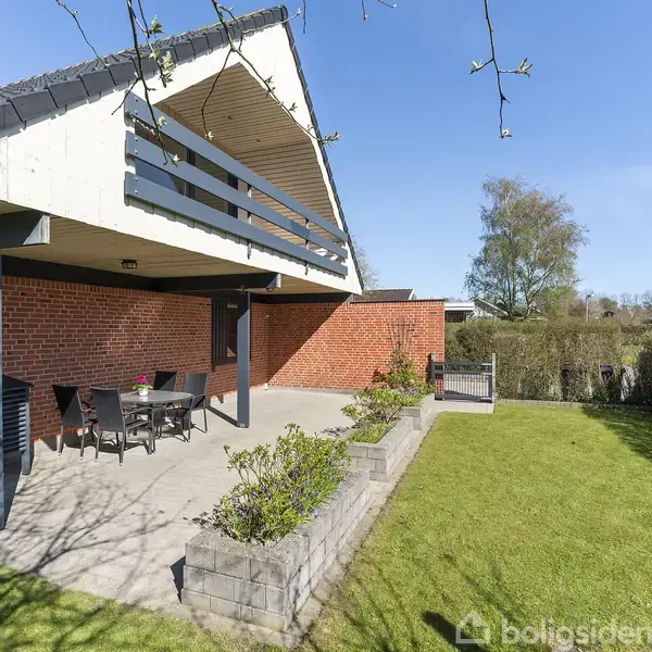 Rød murstensbygning med overdækket terrasse. Terrassen har havemøbler og blomsterbed. Græsplæne omkranser området, og der er hæk mod baggrunden. Klar blå himmel og lidt træer ses i baggrunden.