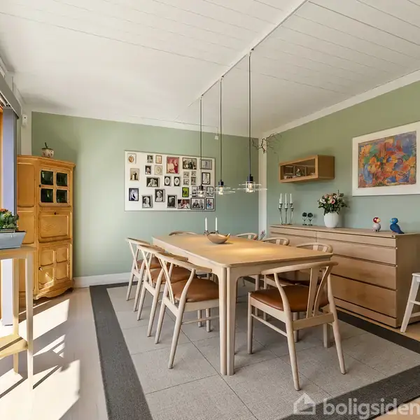 Køkken med hvide skabe og en indbygget ovn, et bord med fire stole. Store vinduer lukker naturligt lys ind. Grønne vægge og trægulve skaber en lys atmosfære.