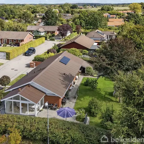 Et rødt murstenshus har en glaskarnap og et solpanel på taget, omgivet af en grøn have med et blåligt parasol. Det ligger i et villakvarter med flere huse.