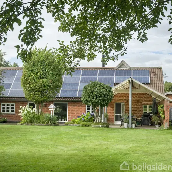 Et rødt murstenshus med solpaneler på taget står i en velplejet have. Træer omgiver huset, og en overdækket veranda ses foran.