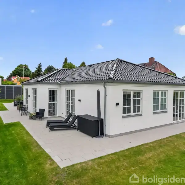 Hvidt hus med sort tag ligger på en grøn græsplæne. Der er terrassemøbler langs husets facade, omgivet af hække og en blå himmel.