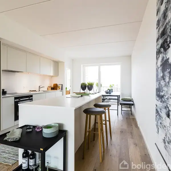 Køkken med hvidt skabssystem og central køkkenø med barstole. Der er en sort/hvid kunstplakat på væggen. Området er lyst og moderne med trægulv og udsigt til terrasse.