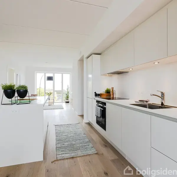 Et moderne køkken med hvide skabe og indbygget ovn strækker sig langs en væg. En køkkenø med planter i potter står midt i rummet. Store vinduer i baggrunden.