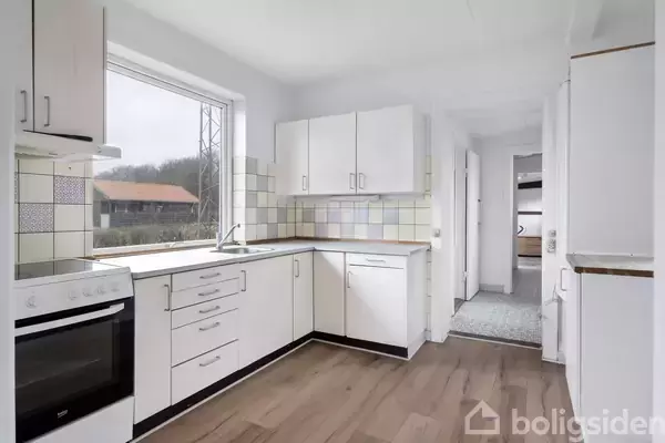 Køkken med hvide skabe og skuffer står stille i et rum med trælaminatgulv og store vinduer. Der er udsigt til et grønt landskab udenfor.