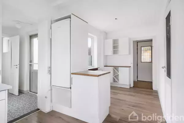 Hvidt, rummeligt køkken med indbyggede skabe og køleskab. Rummet har trælaminatgulv, adskillige døre og et vindue, der lukker lys ind. Til venstre er der en indgang med flisegulv.