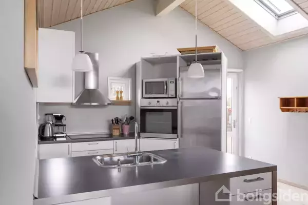 Et moderne køkken med en metalbordplade indeholder en vask, komfur, kaffemaskine og køleskab. To hængende lamper giver belysning, mens et ovenlysvindue lader naturligt lys ind i rummet.