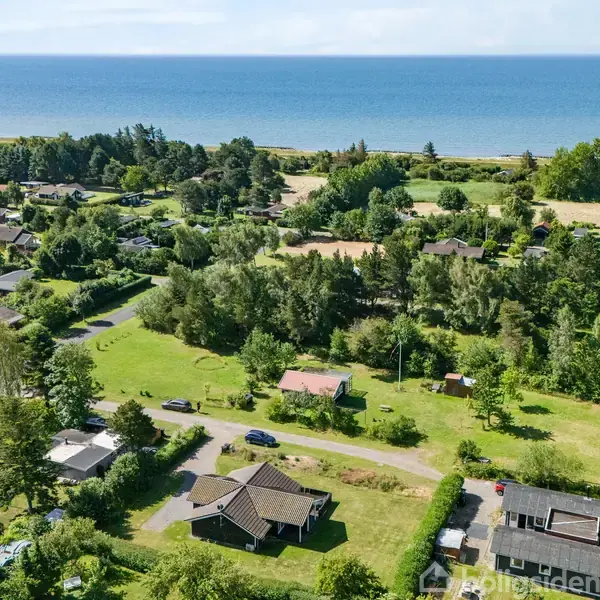 Flere huse ligger spredt i et grønt, skovklædt område nær en kyst. Veje snor sig gennem landskabet, og havet ses i baggrunden mod horisonten.
