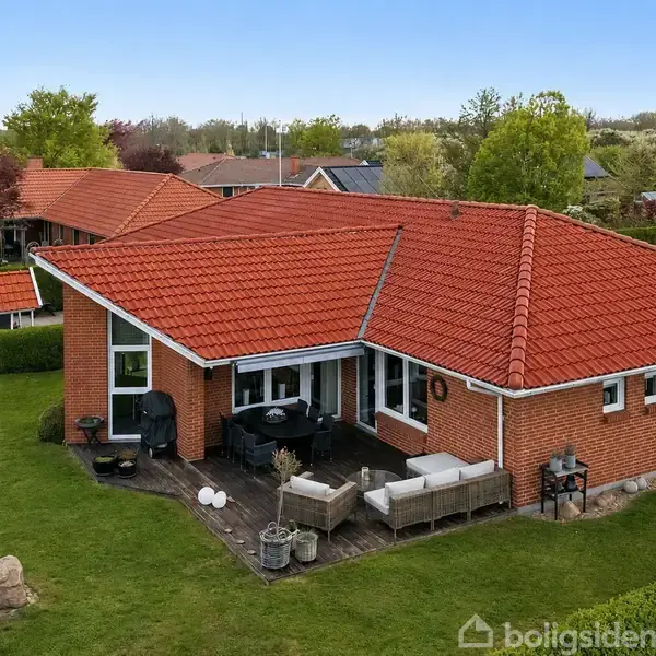 Rødt murstenshus med terracotta tag, omgivet af frodig have og hæk. En terrasse med havemøbler og grill står foran huset. Nabolaget består af lignende boliger og grønne områder.