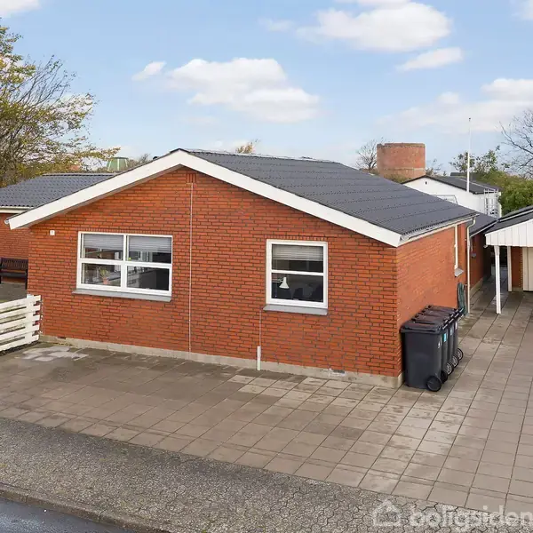 Rødt murstenshus med fladt tag, står stille ved siden af en carport på asfaltbelagt indkørsel, omgivet af et hvidt stakit og grønne træer i forgrunden.