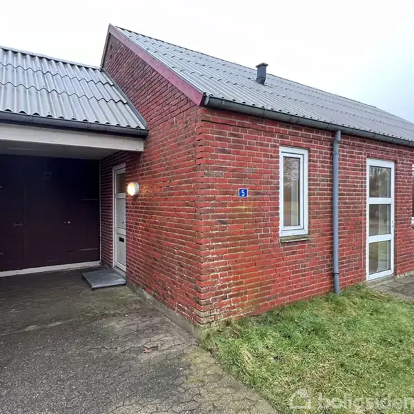 Rødt murstenhus med garage til venstre, belyst af en udendørslampe. Stien og en græsplæne omgiver huset. Nummer 15 er synligt ved indgangen.