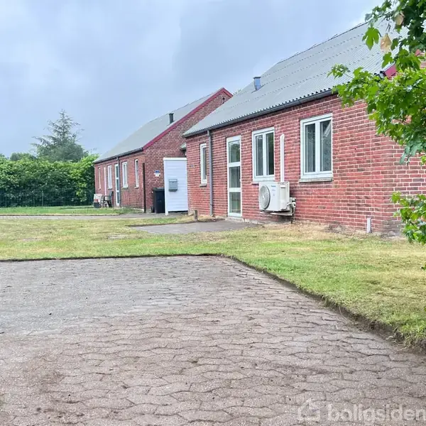 Rødt murstenshus med to hvide vinduer og en havedør, står ved siden af en grusbelagt gårdsplads og græsplæne. En vej og buskads ses i baggrunden.