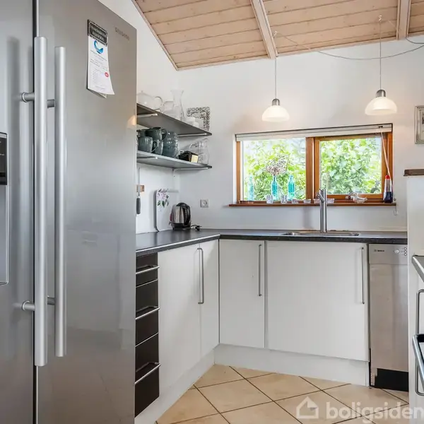 Et moderne køkken med stål-køleskab, hvidt skabsinteriør og indbygget opvaskemaskine. To lamper hænger fra det træbeklædte loft. Et vindue med udsigt til grønne træer lader naturligt lys ind.