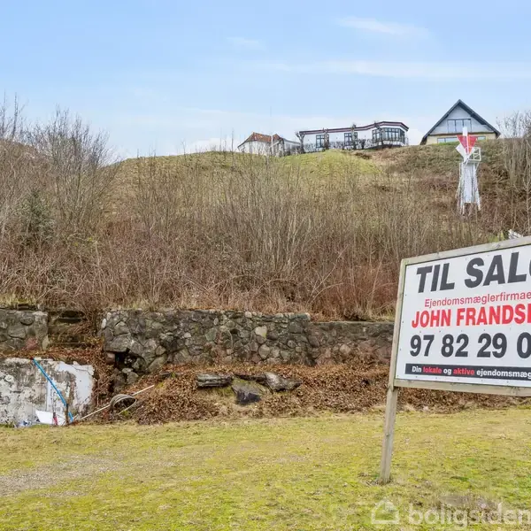 Et skilt står på græsset med teksten "TIL SALG, Ejendomsmæglerfirmaet JOHN FRANDSEN, 97 82 29 00, www.johnfrandsen.dk" foran en bakke med buske og huse i baggrunden.