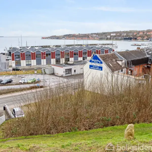 En bygning med et skilt fra "Tømrerfirmaet Mimøller" står blandt flere industrielle strukturer nær en havn. Konteksten er en bakke med græs, buske og udsigt til vandet.
