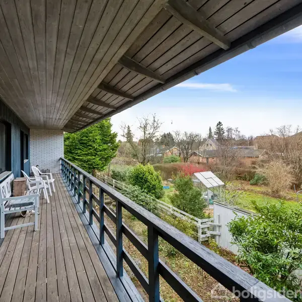 Balkon med træmøbler, der står stille langs en blå husfacade. Udsigt over en grøn have og huse i baggrunden under en lyseblå himmel.