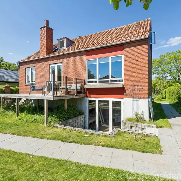 Rødt murstenshus med altan, stående i solrig have med grønne græsplæner og flisegang. Tydelig skorsten og store vinduer præger facaden, mens den blå himmel danner baggrunden.