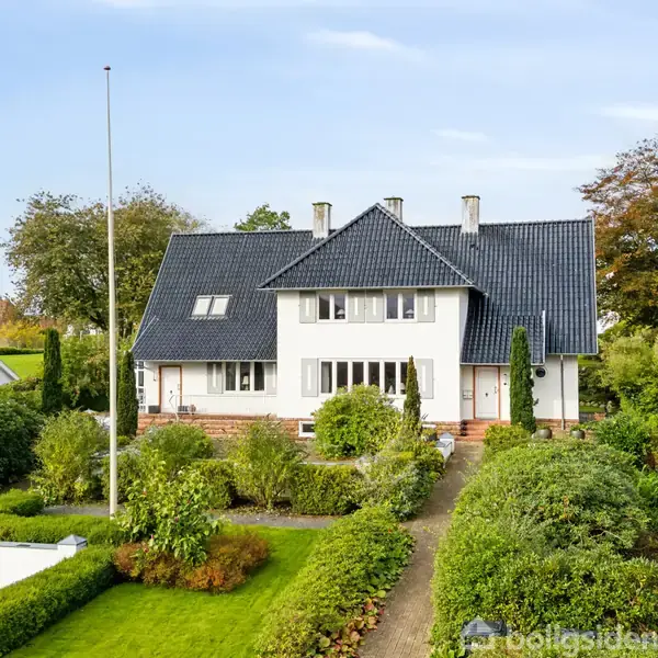 Et hvidt hus med sort tag står omgivet af grøn bevoksning og træer under en klar himmel. Huset har flere vinduer og en indgang med trappe foran.