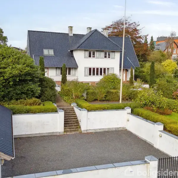 Et hvidt to-etagers hus med blå tagsten står centralt. En grusbelagt gårdsplads er foran huset, der er omgivet af velplejede buske og træer. Baggrunden viser flere moderne bygninger.